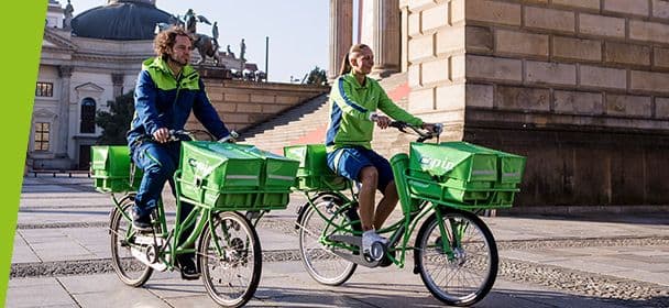 Zwei PIN-Zusteller:Innen fahren nebeneinander auf dem Fahrrad durch Berlin